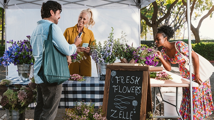 People buying flowers at an outdoor booth with a Zelle accepted here sign.