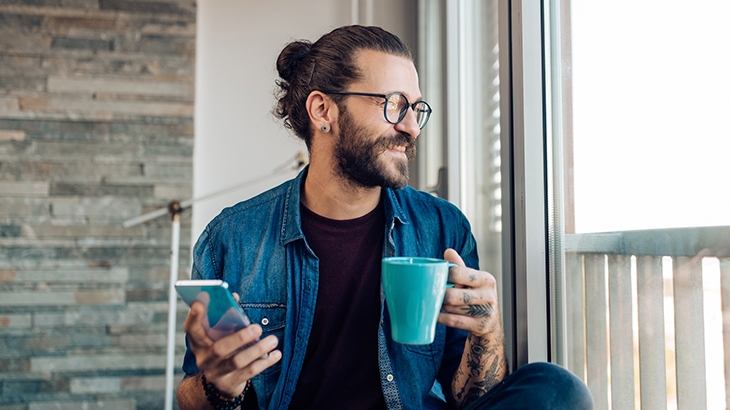 Man relaxing at home, drinking coffee and using a smartphone