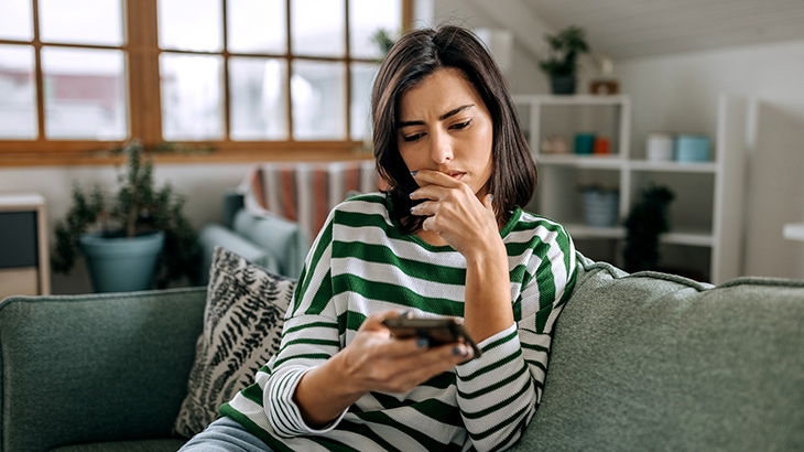 Concerned woman looking at a smartphone sitting on a couch.