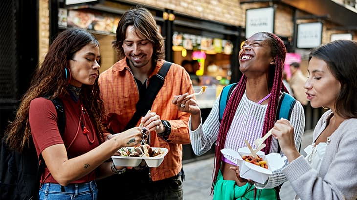 Group of four diverse friends laughing and eating street food outdoors.