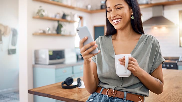 Woman holding a coffee mug and smiling while looking at her smartphone in a kitchen.