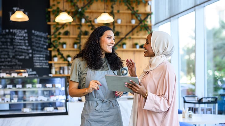 Cafe worker chatting with a customer in a hijab holding a tablet.