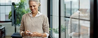 Person standing indoors and looking at a phone, with large windows and greenery in the background.
