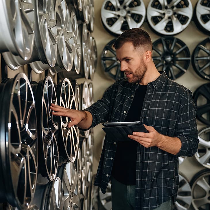 A person in a plaid shirt holding a digital tablet while inspecting a wall of various car wheel rims in a shop.