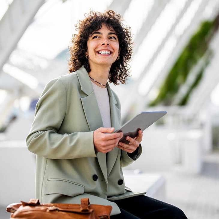 A cheerful woman with dark, curly hair, wearing a light green blazer, sits outside holding a tablet and looking up and away with a wide smile.