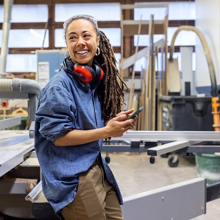 A smiling person with long locs and safety glasses sitting in a woodworking shop holding a smartphone.