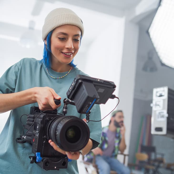 A person with blue hair and a white beanie smiling while operating a professional cinema camera in a bright studio.