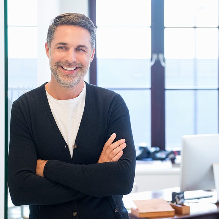 A casually dressed, smiling professional man with salt-and-pepper hair stands with his arms crossed in a modern office.