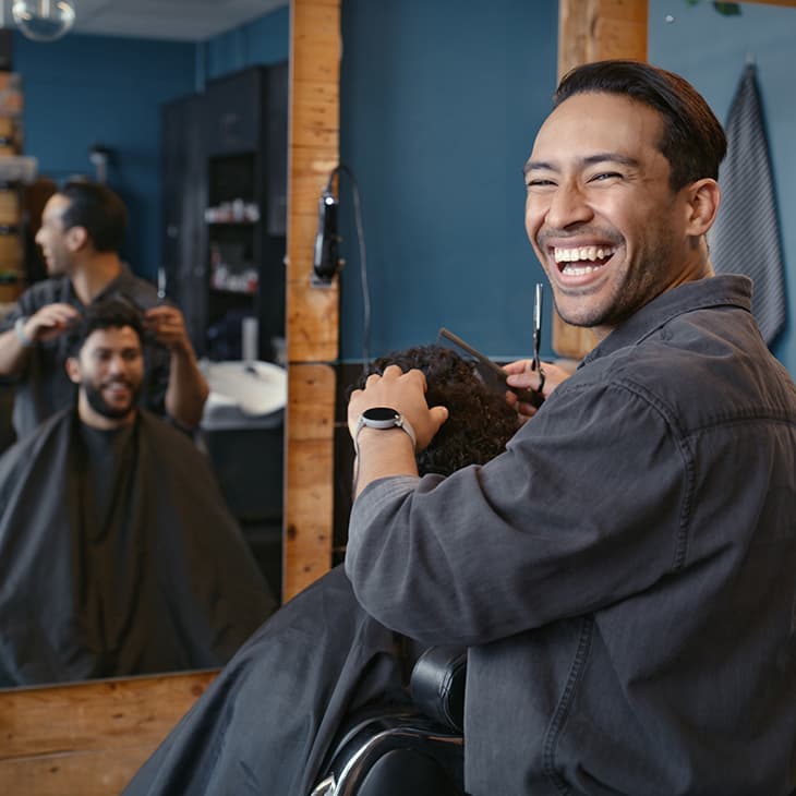 A smiling barber in a grey shirt looks at the camera while cutting a client's hair in a modern barbershop with wood-framed mirrors and blue walls.