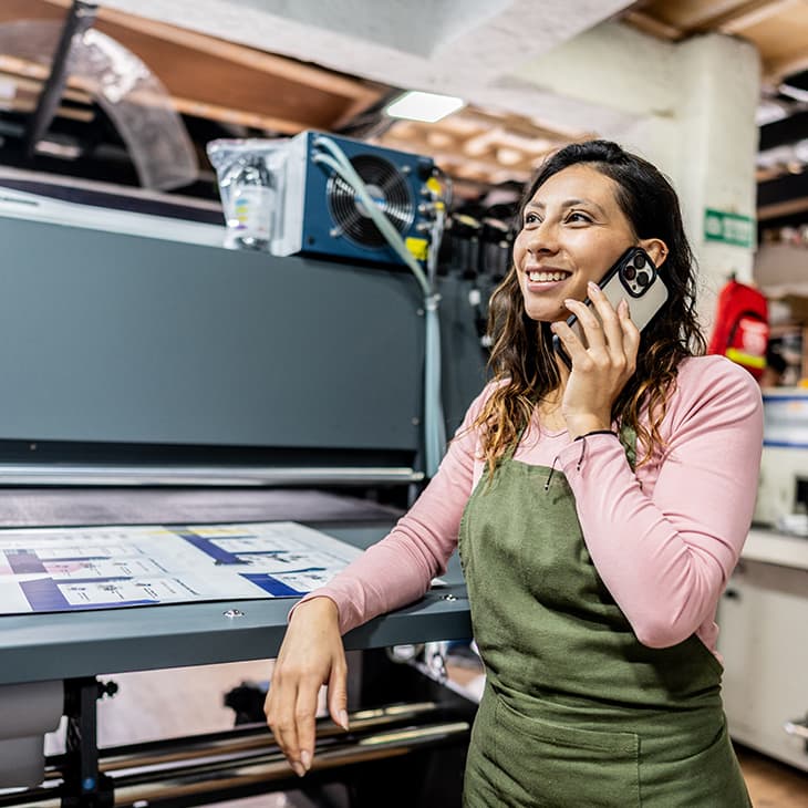 A woman in a green apron smiles while talking on a smartphone in a print shop next to a large industrial printer.