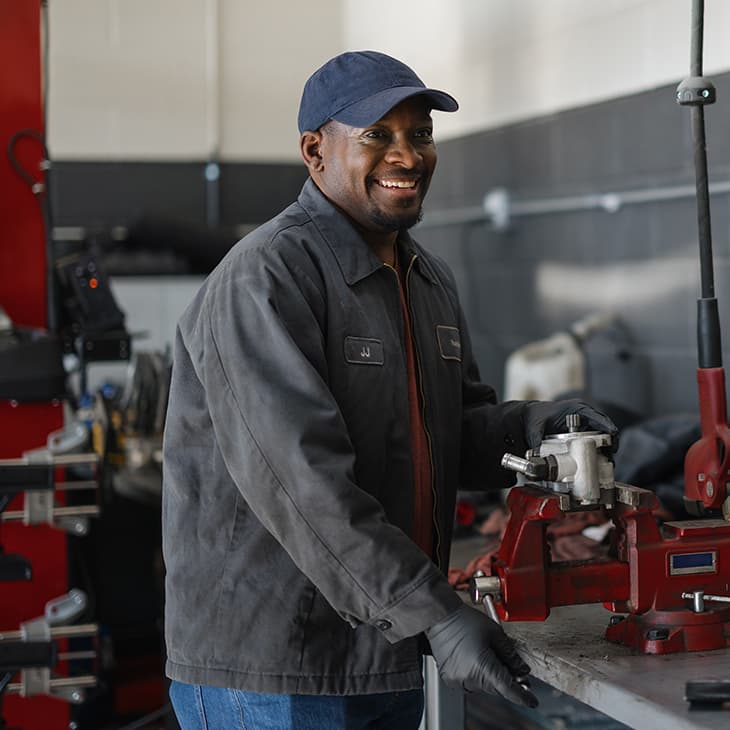 A smiling man in a work jacket and baseball cap works at a workbench in an automotive or machine shop.