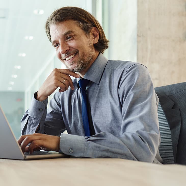 A man in a shirt and tie smiles while working on a laptop at a desk in a bright office.
