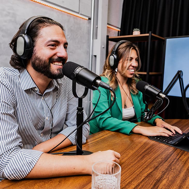 Two people wearing headphones smile and talk into professional microphones while recording a podcast in a studio.