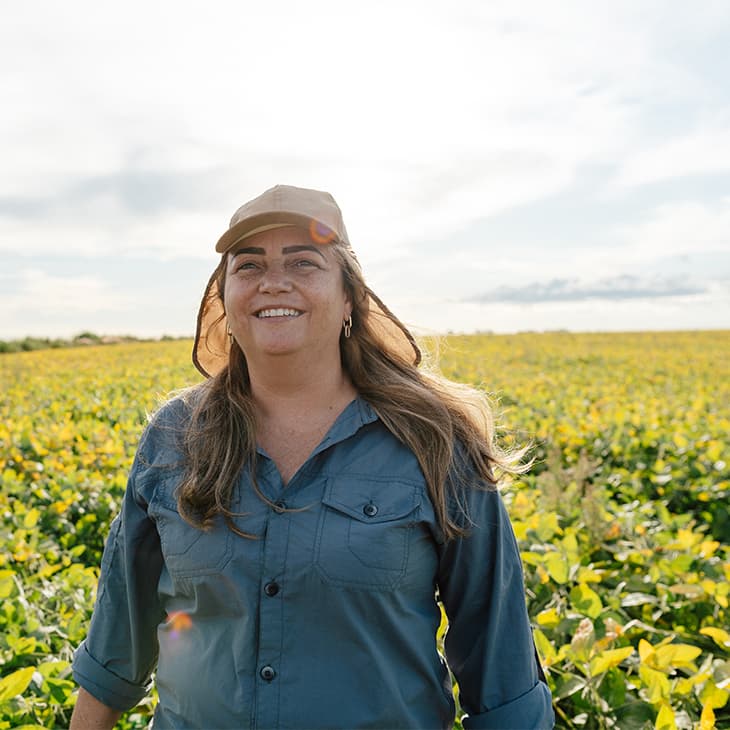 A smiling woman wearing a sun hat and a work shirt stands in the middle of a vast, sunny agricultural field.