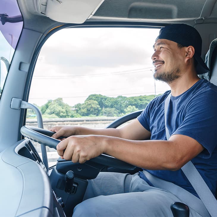 A smiling man in a baseball cap and a t-shirt drives a large commercial truck on a highway.
