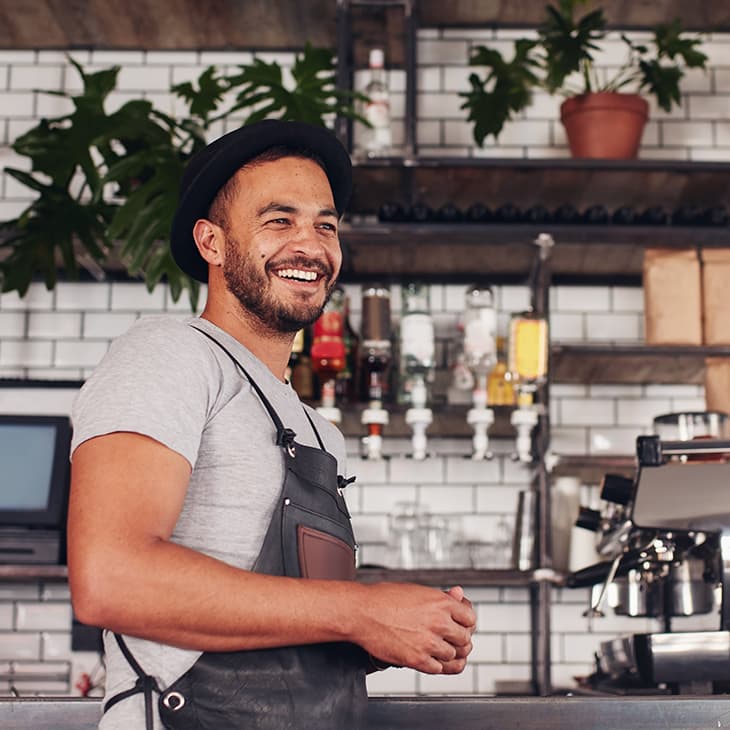 Smiling man in a black apron and fedora standing behind a counter with an espresso machine in a cafe.