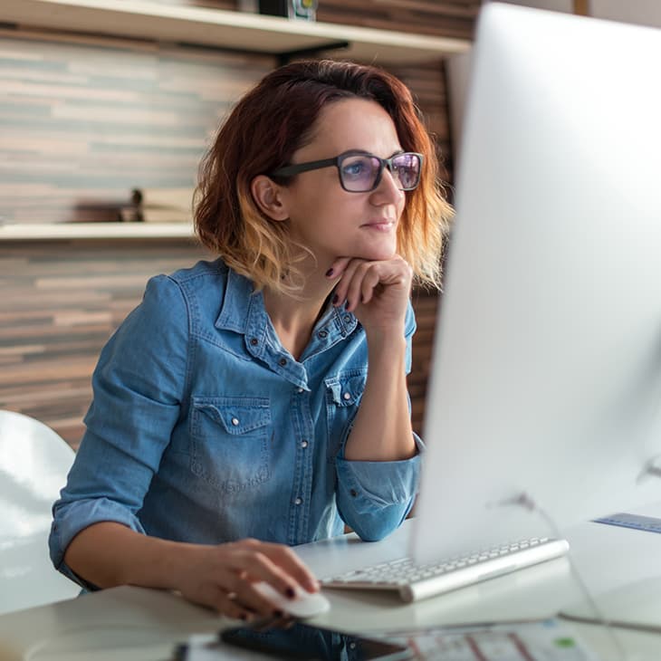 A woman with reddish-brown hair and glasses sits at a desk, looking thoughtfully at a computer monitor in a modern office setting.