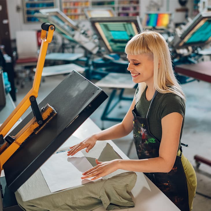 A smiling woman with blonde hair and a dark apron works at a heat press machine in a screen printing studio.