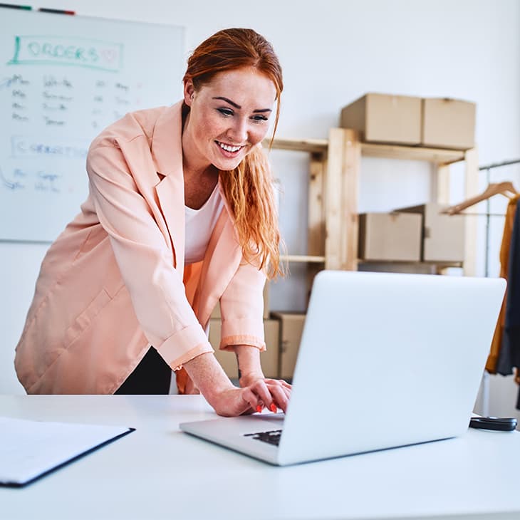 Smiling woman with reddish hair working on a laptop in a small business office surrounded by clothes on a rack and boxes.