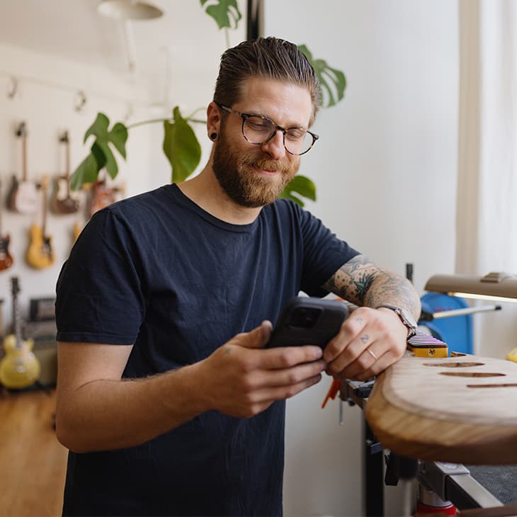 A man with a beard and glasses looks at his smartphone while leaning against a workbench in a guitar workshop.