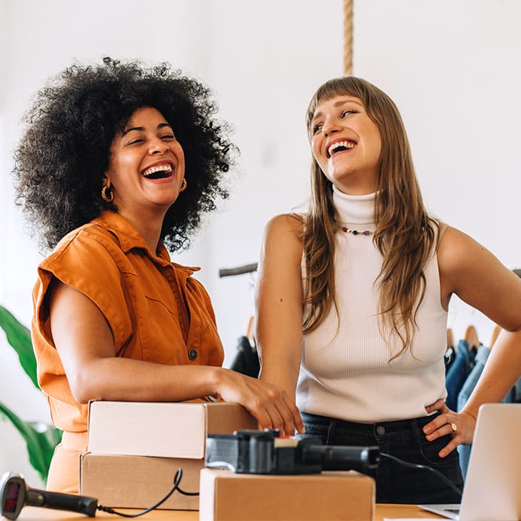 Two women laugh together in a brightly lit clothing boutique while standing near shipping boxes and a laptop.