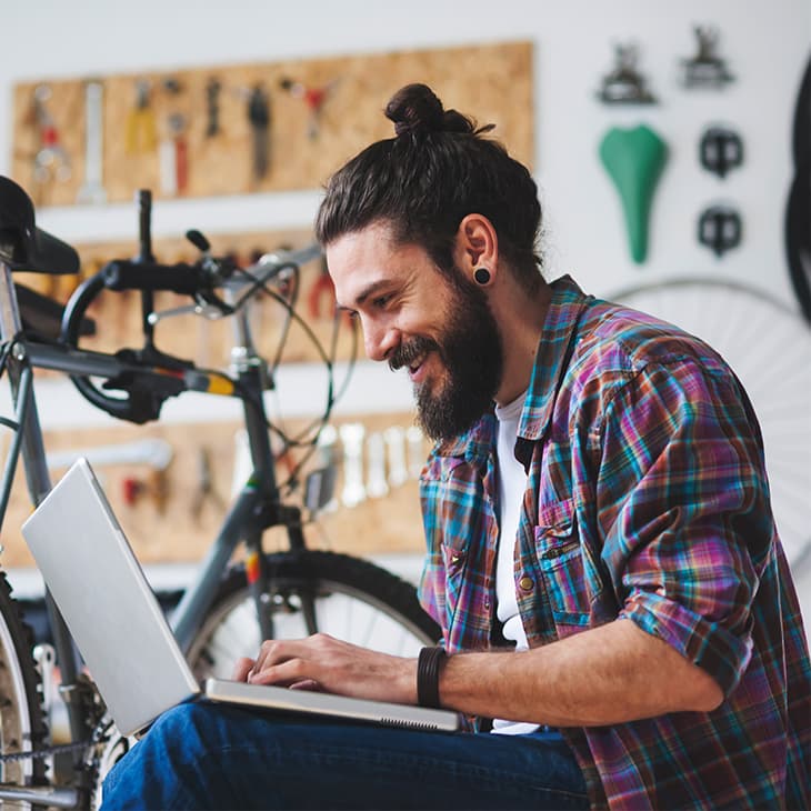 A smiling man with a bun and beard types on a laptop while sitting on the floor of a bicycle repair shop.