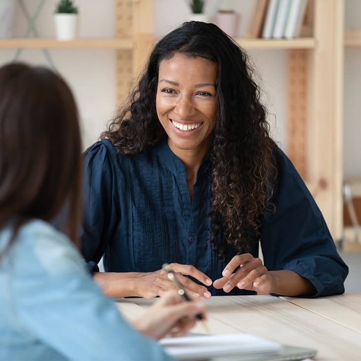 A woman with curly dark hair smiles warmly while sitting across a wooden table from another person during a conversation in a bright office.