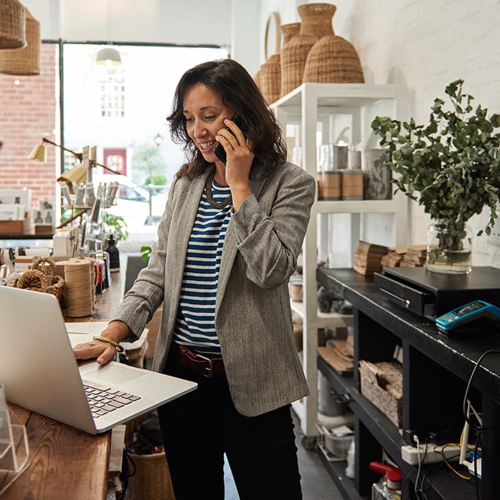 A woman in a grey blazer talks on a cell phone while using a laptop on a wooden counter in a boutique shop filled with woven baskets and home goods.