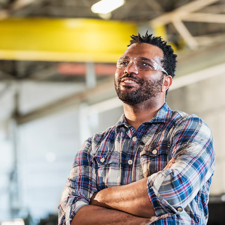 A smiling man in a plaid shirt and safety glasses stands with his arms crossed in a bright industrial workshop.