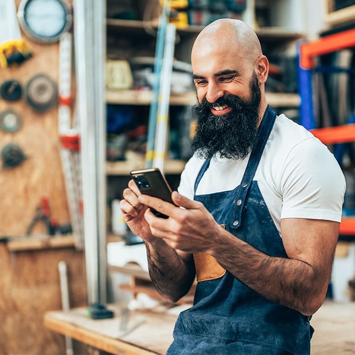 A smiling man with a long beard and denim apron looks at his smartphone in a woodworking workshop.