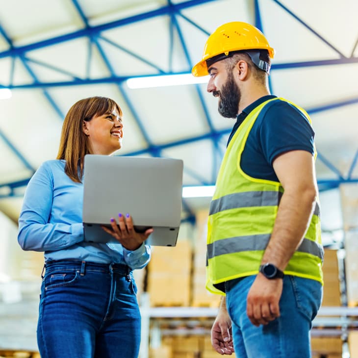 A woman holding a laptop talks with a man wearing a yellow hard hat and safety vest in a large warehouse with high ceilings.