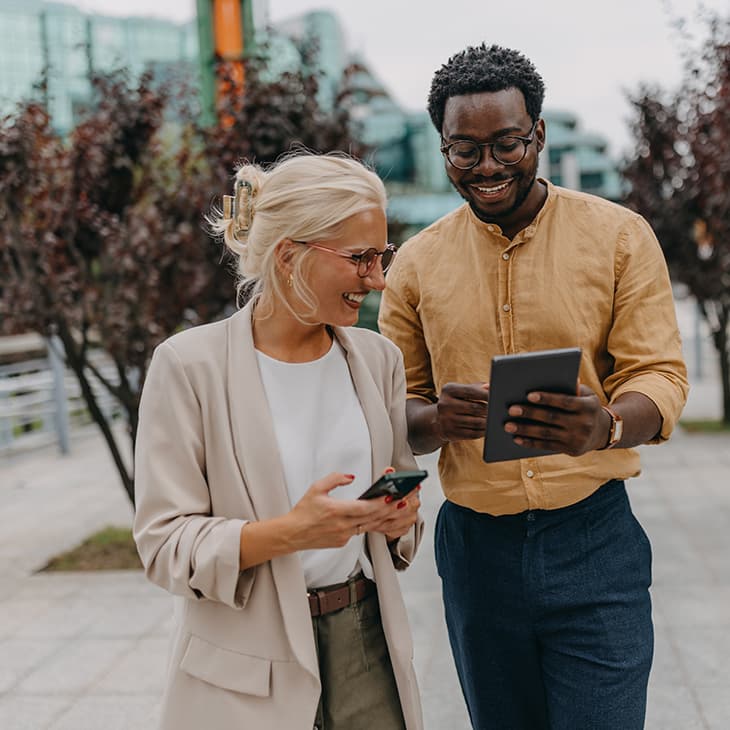 Two smiling professionals standing outdoors, looking at a tablet together.