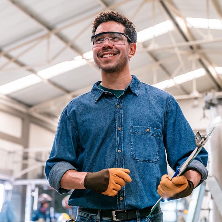 A man wearing safety glasses, a denim shirt, and work gloves smiles while holding a clipboard in an industrial warehouse.