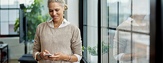 Person standing indoors and looking at a smartphone, with large windows and greenery in the background.