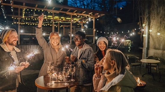 A group of five smiling friends celebrates outdoors at night on a patio decorated with string lights while holding sparklers.