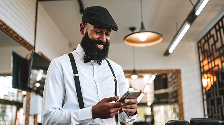 A smiling, well-dressed man with a full beard, wearing a newsboy cap, bow tie, and suspenders, looks at his mobile phone in what appears to be a barbershop or retail space.
