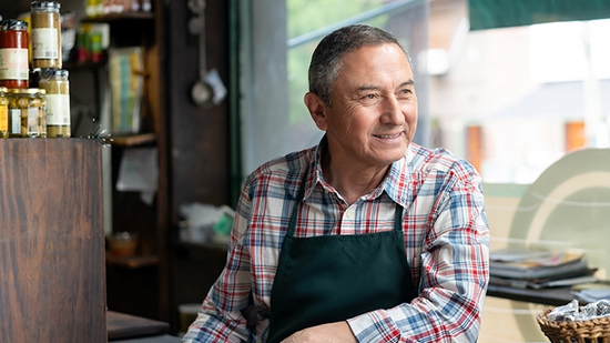 Middle-aged man in a plaid shirt and dark apron smiling and looking out the window of his small business.