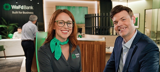 A smiling woman in a WaFd Bank uniform and a man in a blue suit sitting together inside a modern WaFd Bank branch.