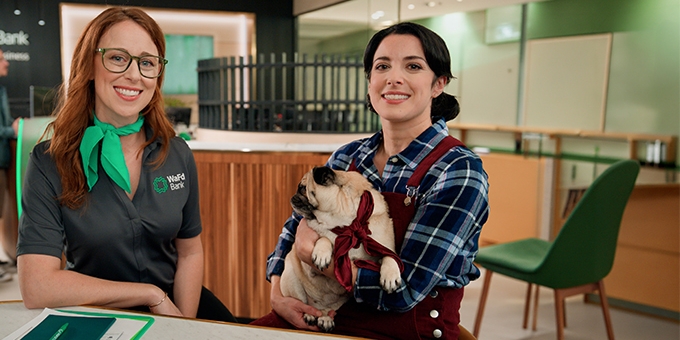 A WaFd Bank employee and a woman holding a pug sit together at a table inside a bank branch.