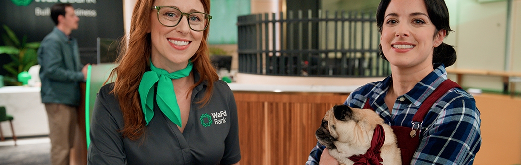 A WaFd Bank employee and a woman holding a pug sit together at a table inside a bank branch.