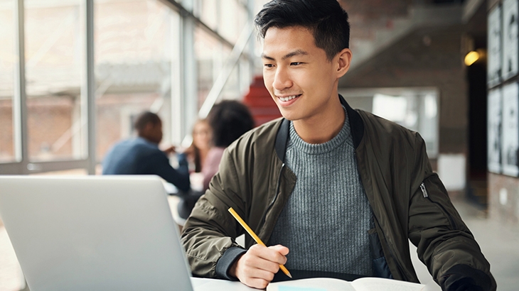 College student studying with laptop and writing in notebook on campus.