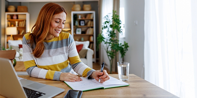 Woman seated at a dining table, planning her time with a notebook and laptop.
