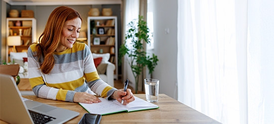 Woman seated at a dining table, planning her time with a notebook and laptop.