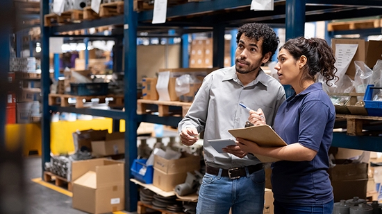 Two colleagues discuss inventory while looking at a tablet and a clipboard in a large warehouse with tall industrial shelving.