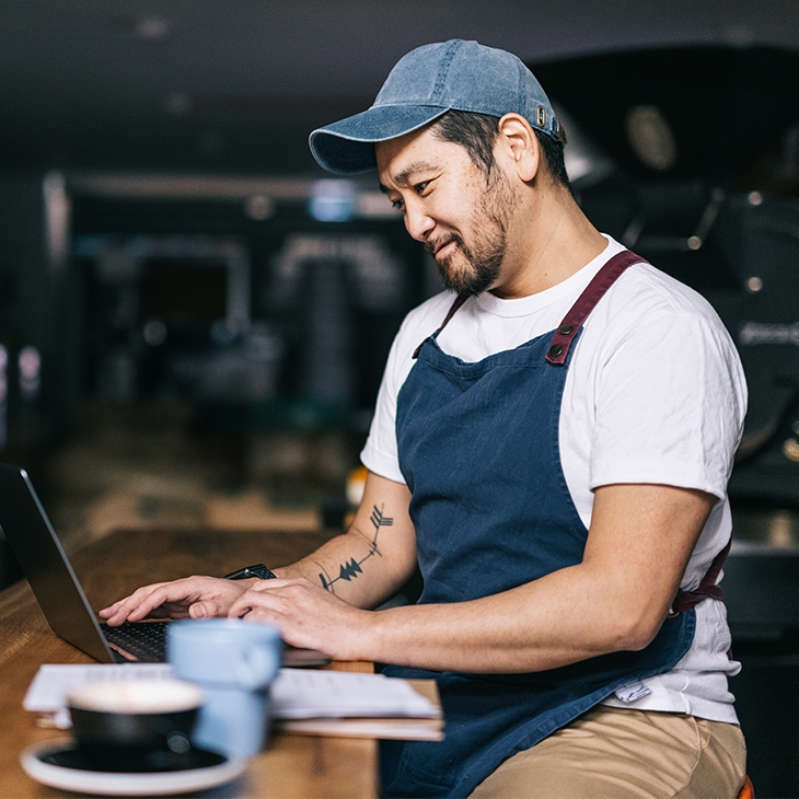 A business owner wearing an apron and a baseball cap, working on a laptop.