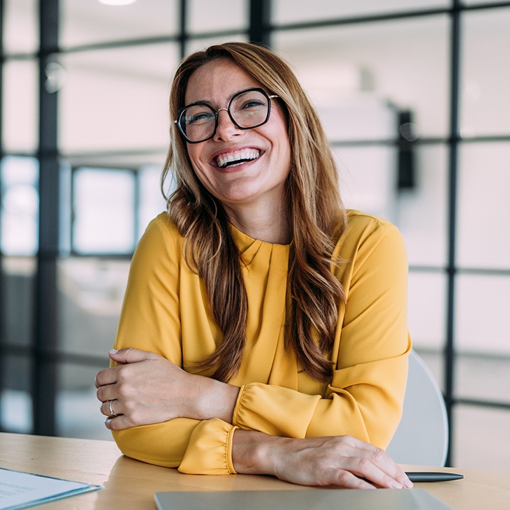 A blonde woman with glasses in a yellow top is sitting at a desk and smiling broadly.
