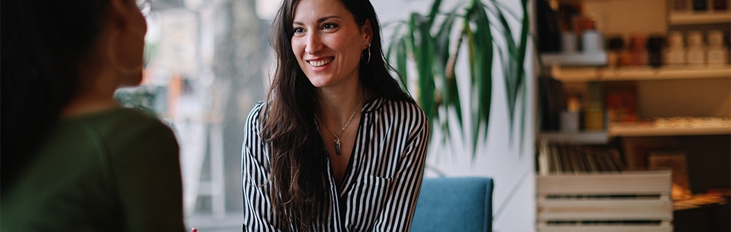 A woman in a striped shirt smiles while holding a tablet and talking to a colleague in a bright, modern cafe setting.