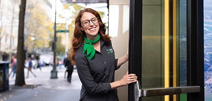 A smiling woman in a gray WaFd Bank uniform and green scarf holds open a glass door to a bank branch.