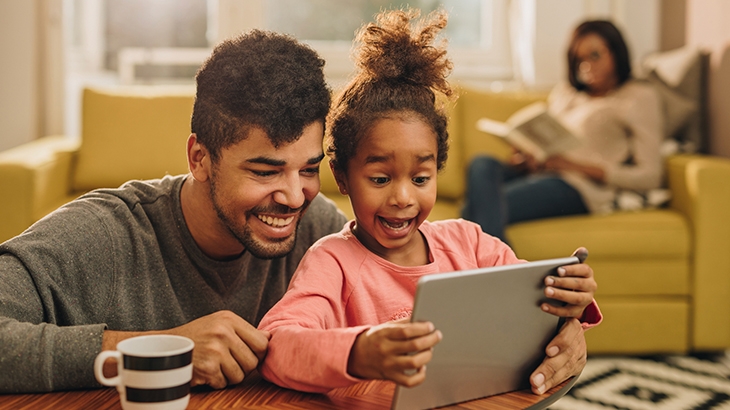 Happy girl using tablet with her father, mother in the background reading on the couch.