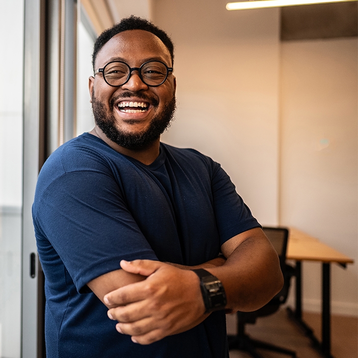 Happy man in a blue shirt and glasses, standing by a window with his arms crossed in an office.